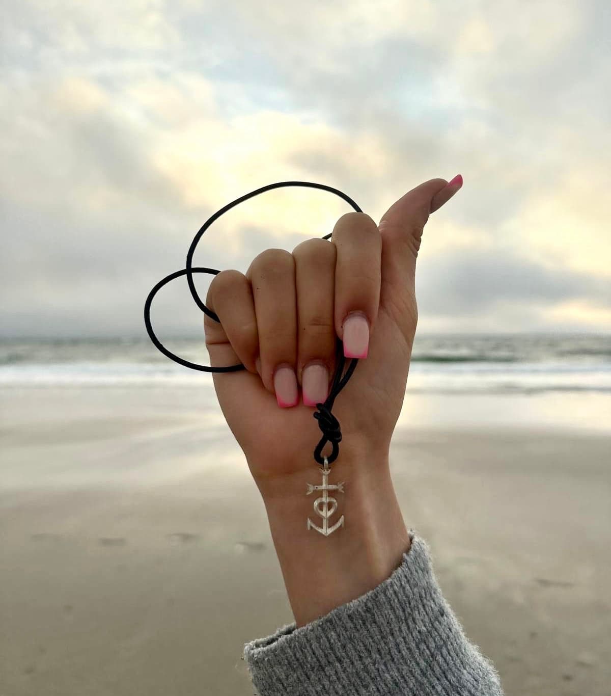 Hand mir rosa lackierten Nägeln  halt Anhänger mit Liebe-Glaube-Hoffnung aus Silber vor Meereskulisse am Strand hoch. 