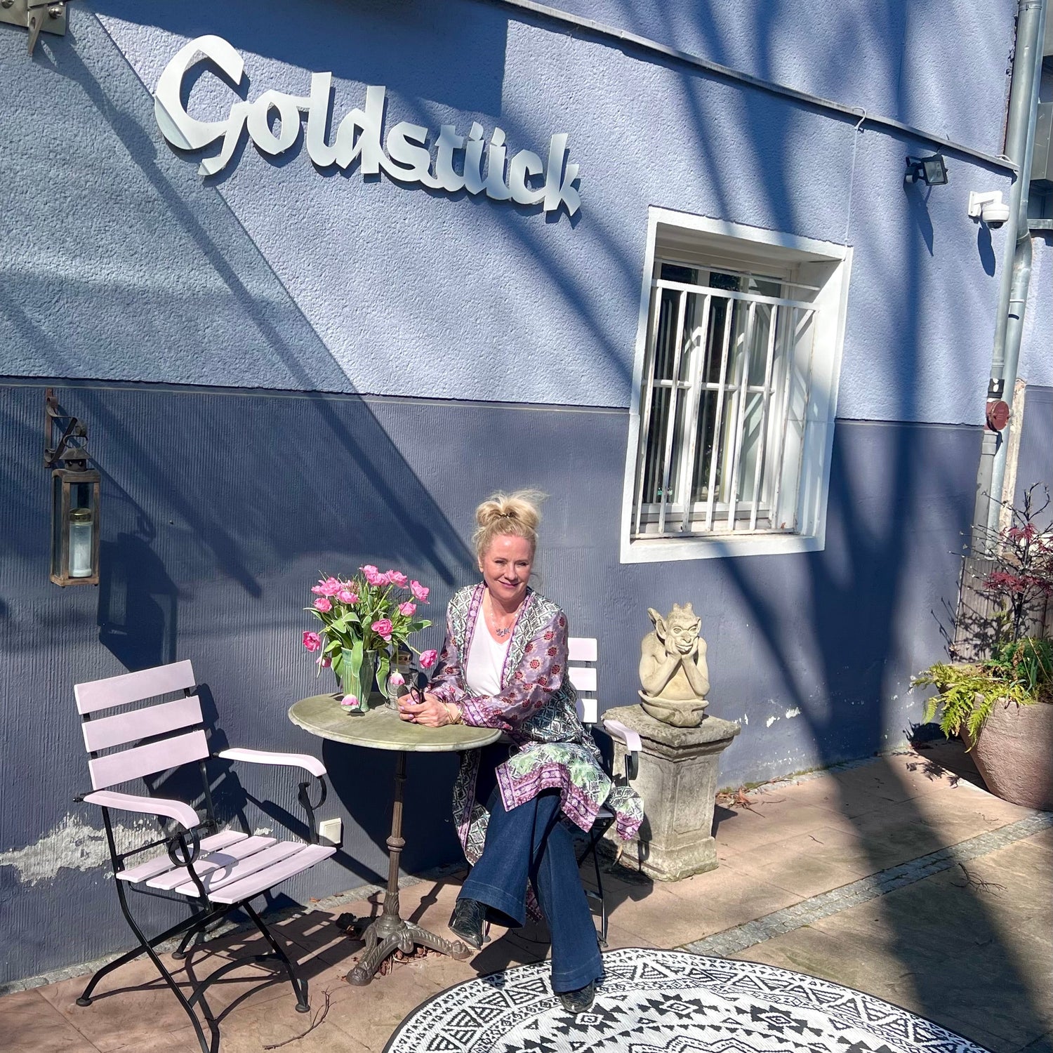 Außenansicht Atelier Q Goldschmiede in Köln mit blauer Fassade, Sitzgelegenheit und Blumendekoration und Andrea Quast vor dem Laden.