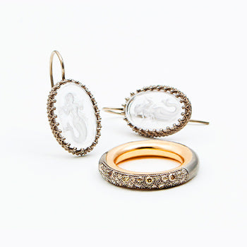 Handgeschmiedete Ringe aus Gold mit Diamanten in Champagnertönen – Ohrschmuck mit Meerjungfrauen-Gravur im Hintergrund